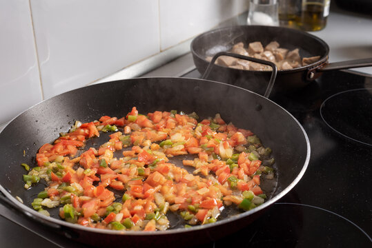 Pan for paellas with poached vegetables, in the background another pan with meat on the hob in the house, cooking at home.