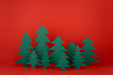 Obraz premium Traditional Christmas festive background with cartoon paper spruces as forest on red backdrop in elegant baby style. New Year mockup for advertising, design, card, poster, flyer.