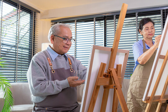 Couple Senior drawing at art. Side view portrait of white haired senior woman holding palette painting pictures at easel in art studio.