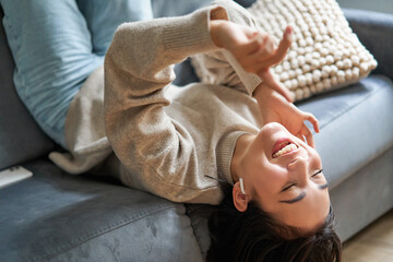 Portrait of happy young asian woman feeling upbeat, lying on sofa and listening music in wireless headphones, smiling pleased - obrazy, fototapety, plakaty