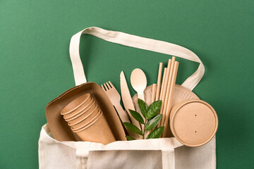 Many paper cups, food containers, paper drinking straws and bamboo cutlery set in white cotton bag over green background. Ecological paper utensils bundle