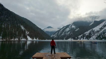 The guy standing on the pier admires the mountain lake. There is a view of the mirrored black color of the water, which reflects snowy mountains, green forest, clouds and a yellow sunset. Kolsai Lake - Powered by Adobe