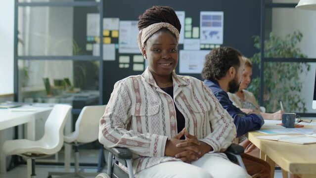 Medium long shot of African American businesswoman in wheelchair posing for camera with smile during workday in office