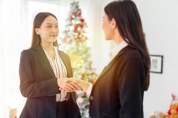 Two young smart beautiful asian lady businesswomen smiling and have a hand shake to commemorate a deal successful in a meeting room in front a Christmas tree
