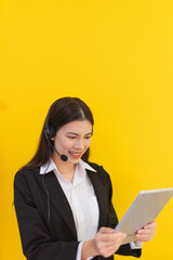 Portrait photo of a young beautiful smart asian lady customer support agent representative with a digital tablet and her wired microphone headset on a yellow color background