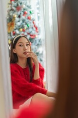 Portrait photo of a young beautiful friendly asian female lady in red long sleeve sweater and white skirt with nice headband posing different looks in front mirror with a nice decorated Christmas tree