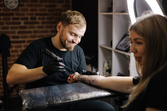 Young smiled tattoo artist is tattooing a woman's hand. Wireless tattoo machine, safety and hygiene at work. Close-up of tattoo artist work. Tattoo salon