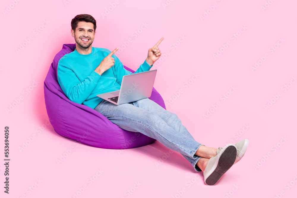 Poster full size photo of nice young man hold netbook sit beanbag point empty space dressed stylish blue ou