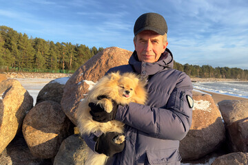 Portrait of happy handsome retired senior man is holding on hands, walking with Pomeranian Spitz dog, cute puppy at winter cold day, sunny weather.