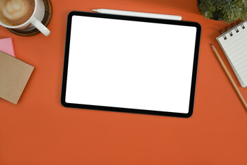 Flat lay digital tablet with white empty screen, notepad, coffee cup and potted plant on orange background