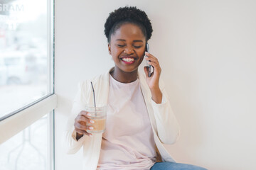 Healthy young businesswoman with glass of fresh beverage, speaking on mobile phone while consulting client sitting in cafe. Cute lady having phone conversation