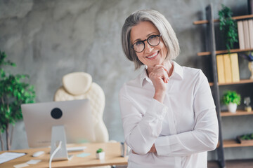 Photo of cheerful experienced company founder toothy smile arm touch chin spacious office inside