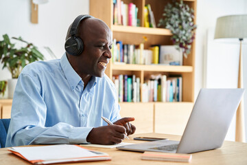 Middle-aged African-American man wearing headphones and taking notes while attending remote conference call while working at home with laptop computer