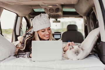 Woman with a bobble hat Using Laptop While Lying With cat In Camper Trailer. Digital nomad lifestyle - obrazy, fototapety, plakaty