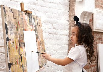 Obraz na płótnie Canvas Dreamy pretty kid girl with paintbrush near drawing easel. Little painting artist.