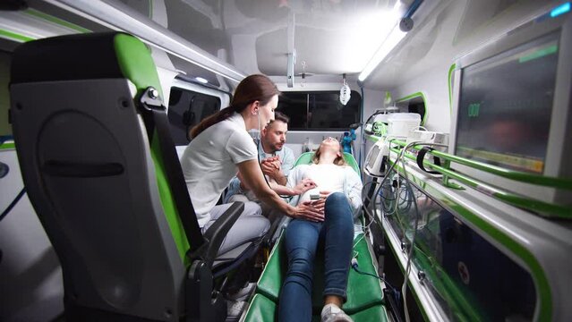 Young caucasian woman lying on stretchers in ambulance during labor. Professional female paramedic measuring heartbeat of baby with stethoscope providing help patient holding hand of husband.