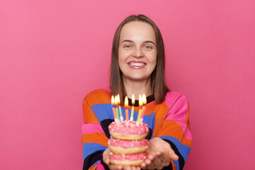 Portrait of satisfied brown haired woman holding out delicious appetizing doughnuts with burning candles, wearing jumper, standing isolated over pink background, looking at camera with toothy smile.