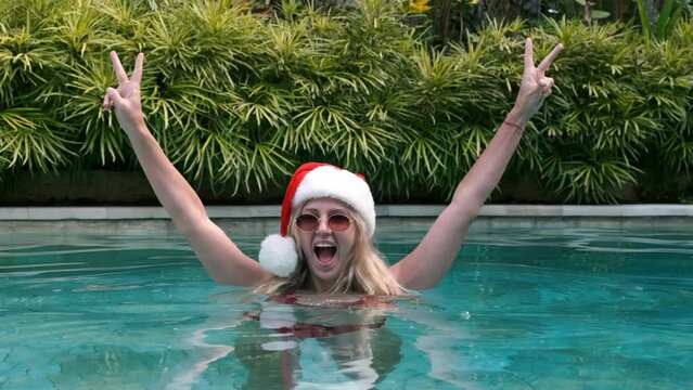 Joyful young blonde woman wears Santa's hat, red bikini and sunglasses celebrating Christmas in pool against green plants on vacation. Girl screams yeah raising her hands showing two fingers.