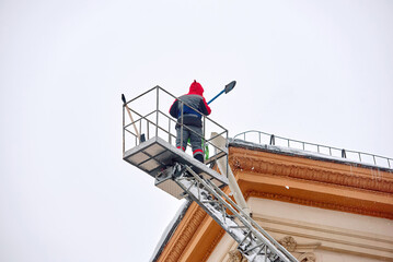 Naklejka premium Man on lift crane remove icicles and snow from roof. Man with shovel breaking ice and hit icicles on rooftop, clearing snow from roof of an old building. Worker in crane bucket remove ice at height