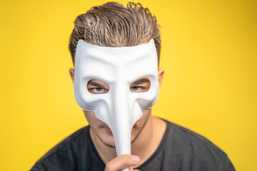 Handsome young man expressive eyes covers his face masquerade white theatrical mask with long nose yellow background. Gigalo alphonse model cheating seducer. Hiding emotions psychological masks.  - obrazy, fototapety, plakaty
