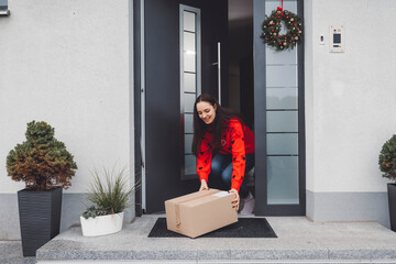 Young woman in red Christmas sweater picking up a cardboard box mailman left out at the front door