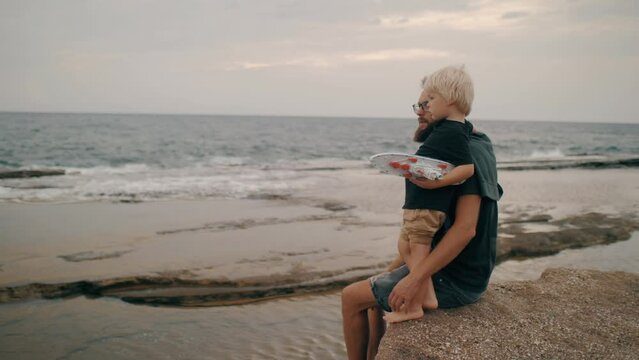 Young bearded man enjoy parenthood hugging cute little boy talking at sea wave coastline