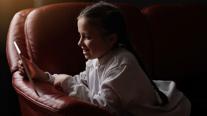 Curious cute teen girl using digital tablet technology device sitting on sofa. Little child holding pad computer surfing internet play game at home. Children tech addiction concept. Happy childhood © Andriy Medvediuk