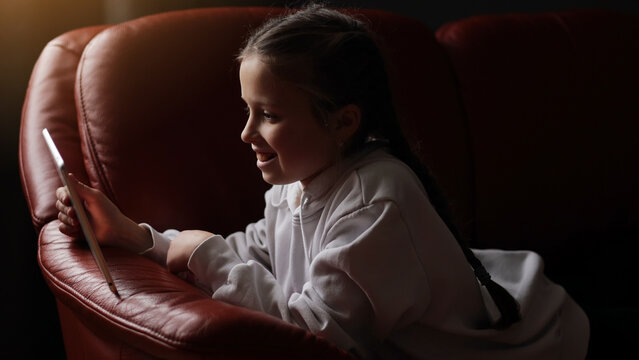 Curious cute teen girl using digital tablet technology device sitting on sofa. Little child holding pad computer surfing internet play game at home. Children tech addiction concept. Happy childhood