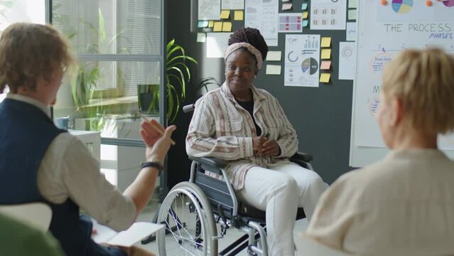 African American female business coach with disability sitting in wheelchair and answering questions about scheme on flipchart while teaching employees in office