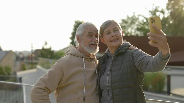 Happy senior couple in love doing selfie standing in the park. Elderly family spending time together, using smartphone. Mature people relationship and technology concept.