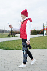 Woman wearing knee brace or orthosis after leg surgery walking in the park using smartphone