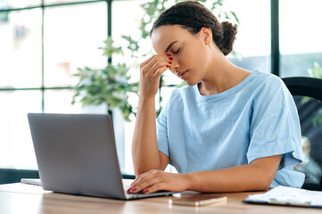 Exhausted stressed brazilian or hispanic young woman, office employee, sits at the work desk in the office, overworked, feeling tired of working in a laptop, having a headache, migraine, needs a break