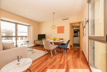 A living room with round glass dining table with glasses of orange juice with velvet upholstered chairs in various colors and reddish wooden floors
