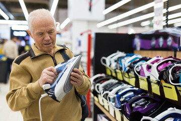 elderly grayhaired man pensioner looking iron at counter in showroom of electrical appliance...