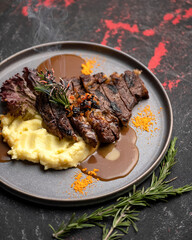Beefsteak garnished with mashed potatoes and sauce on dark metal plate. Decorated with sprig of rosemary. Grill menu. Grilled meat. Serving the dish. View from above. Soft focus. Dark background. 