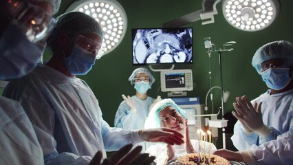 Portrait of young happy female patient celebrating birthday blowing out candles in operation room with multiethnic group of surgeons and nurses. Cheerful doctors applauding smiling. - Powered by Adobe