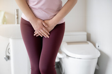 A woman wearing leggings is complaining of pain from urinary incontinence in front of the toilet - obrazy, fototapety, plakaty