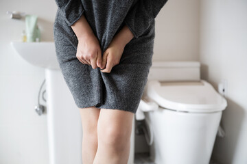 A woman wearing knitwear is complaining of pain from urinary incontinence in front of the toilet - obrazy, fototapety, plakaty