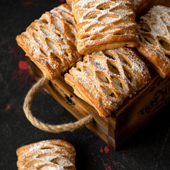 Homemade baking. Puff pastry with fruit filling for family tea drinking. The pie is sprinkled with powdered sugar, laid out on wooden board. Suitcase with pastries.Dark background. Soft focus.