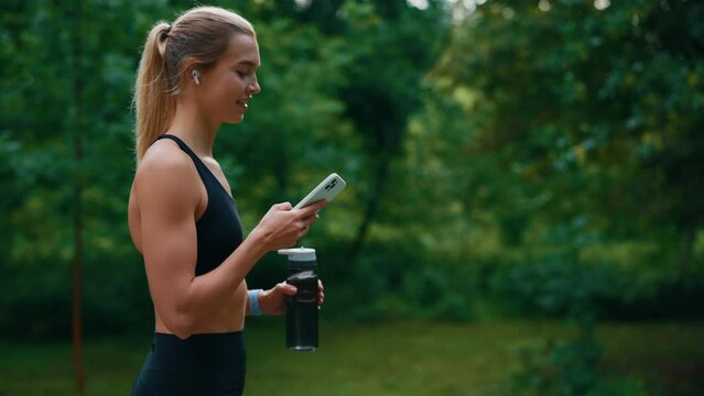 Side view of the sporty woman walking, holding sport bottle using, scrolling phone and listening to music using earphones. Sport and technology and healthy lifestyle concept.