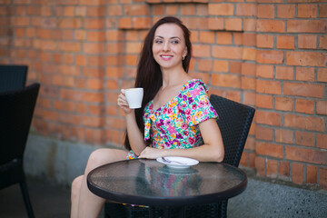 Stylish slender smiling woman with long dark hair, with a cup of coffee, in a light summer dress with a floral pattern, sits at a table in a cafe on the street, against the backdrop of a brick wall.