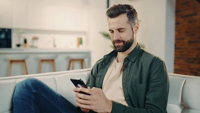 Positive handsome stylish caucasian man sits on a sofa in a cozy living room, uses his smartphone, spends leisure time online, chats with friends on social networks, browsing the internet, smiles