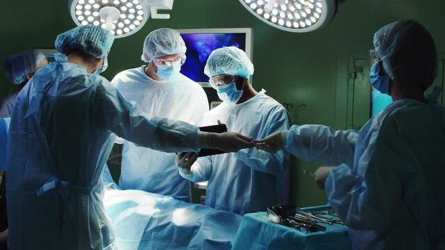 Group of surgeons performing operation in hospital using tablet and instruments. Professional medical workers in medical gown, mask and cap. Multiethnic qualified doctors operating in operation room.