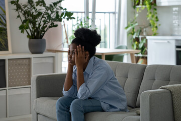 Uninterested discouraged African woman sits on couch at home not in mood clutching head looking into distance. Bored young black girl is sad after quarrel with friends or boyfriend's departure