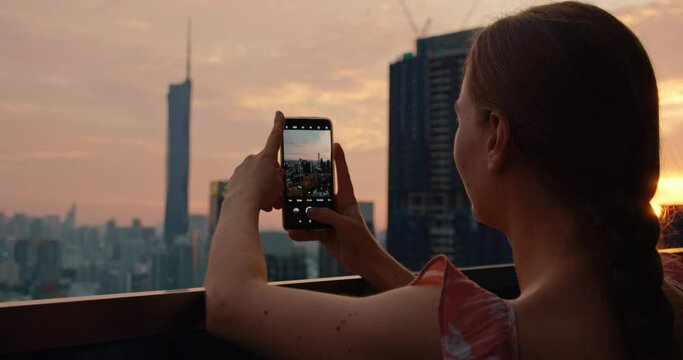 Rear view of woman holds a phone in hands and takes a photo of evening sunset city Kuala Lumpur Malaysia. Outdoors on terrace with modern high buildings. Go everywhere traveling concept.