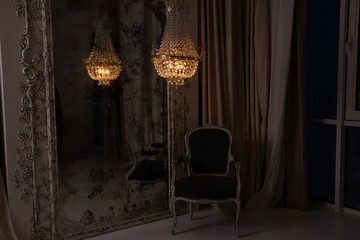 A crystal old chandelier hangs near the mirror and there is a chair next to the house in the...
