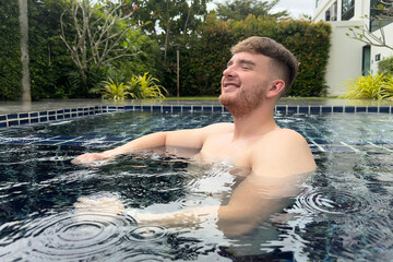 Happy man, young positive guy enjoying summer day, vacation in swimming pool in the yard of house, smiling