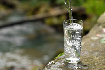 Fresh water pouring into glass on stone near river. Space for text