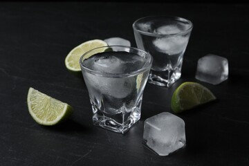 Shot glasses of vodka with lime slices and ice on black table