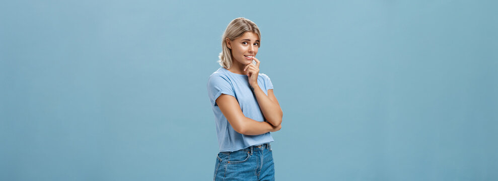 Studio shot of unconfident silly and insecure cute feminine girl with blond hair standing half-turned over blue background holding finger on lip while being shy ask question smiling hesitating